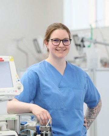 Eine Krankenschwester in blauer Arbeitskleidung steht lächelnd neben medizinischen Geräten in einem modernen Krankenhausumfeld.