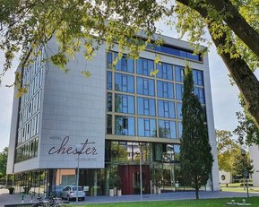Modernes Hotelgebäude mit der Aufschrift "Hotel Chester" in einer grünen Umgebung, umgeben von Bäumen und einem klaren Himmel.