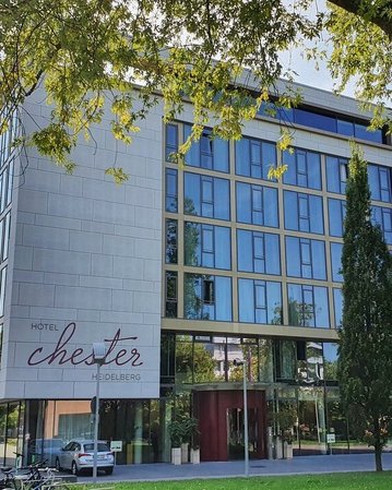 Modernes Hotelgebäude mit der Aufschrift "Hotel Chester" in einer grünen Umgebung, umgeben von Bäumen und einem klaren Himmel.