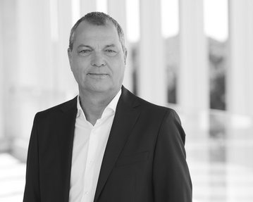 Mann in Anzug mit kurzem Haar, steht vor einer hellen, modernen Architektur. Neutraler Gesichtsausdruck, professionelles Auftreten.