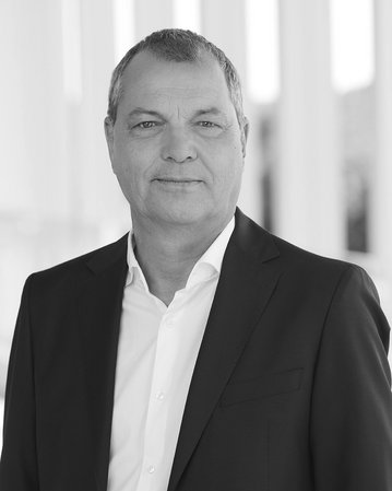 Mann in Anzug mit kurzem Haar, steht vor einer hellen, modernen Architektur. Neutraler Gesichtsausdruck, professionelles Auftreten.