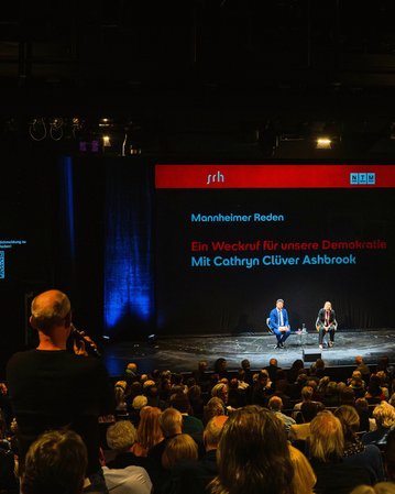 Veranstaltung mit Cathryn Clüver Ashbrook auf der Bühne, Publikum im Vordergrund, Hintergrund mit Text über Demokratie.