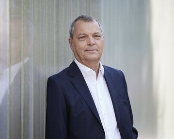Prof. Dr. Christof Hettich trägt einen dunklen Anzug, steht an einer transparenten Wand und schaut direkt in die Kamera.