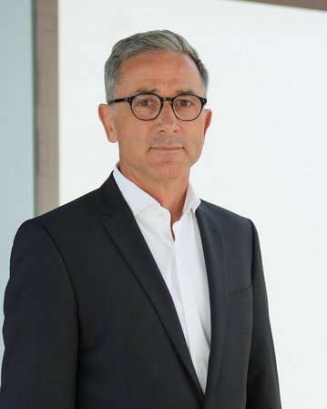 Mann in Anzug mit Brille steht vor einer modernen Glaswand. Er hat graue Haare und schaut direkt in die Kamera.