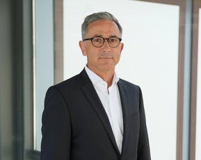 Mann in Anzug mit Brille steht vor einer modernen Glaswand. Er hat graue Haare und schaut direkt in die Kamera.