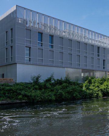 Moderne Gebäude entlang eines Gewässers, mit einer Mischung aus Glas- und Metallfassaden, umgeben von grüner Vegetation.