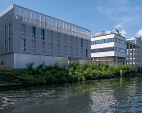 Moderne Gebäude entlang eines Gewässers, mit einer Mischung aus Glas- und Metallfassaden, umgeben von grüner Vegetation.