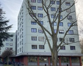 Mehrstöckiges Wohngebäude mit großen Fenstern, umgeben von Bäumen und Fahrrädern im Vordergrund.