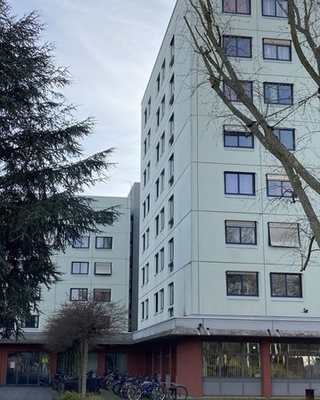 Mehrstöckiges Wohngebäude mit großen Fenstern, umgeben von Bäumen und Fahrrädern im Vordergrund.