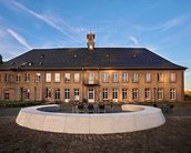 Historisches Gebäude mit einer Fassade aus Sandstein, großen Fenstern und einem Uhrenturm, umgeben von einem runden Sitzbereich.