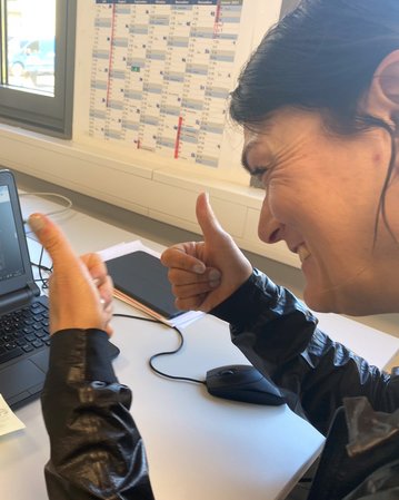 Eine Person sitzt an einem Tisch und zeigt Daumen hoch, während sie an einem Videoanruf auf einem Laptop teilnimmt. Auf dem Bildschirm sind mehrere Teilnehmer zu sehen.