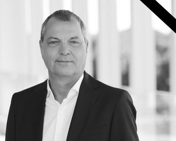 Ein Mann in einem Anzug steht vor einer modernen Architektur, umgeben von Säulen, mit einem Trauerschleier in der oberen Ecke.