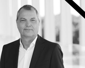 Ein Mann in einem Anzug steht vor einer modernen Architektur, umgeben von Säulen, mit einem Trauerschleier in der oberen Ecke.