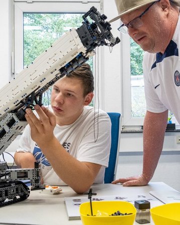 Ein Jugendlicher arbeitet an einem LEGO-Modell eines Krans, während ein Erwachsener ihm zuschaut und Anweisungen gibt.