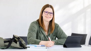 Eine lächelnde Frau mit Brille sitzt an einem Tisch, schreibt mit einem Stift und hat ein Tablet sowie Bücher vor sich.