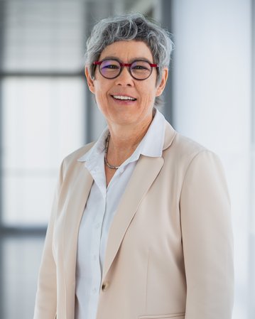 Eine lächelnde Frau mit grauen, lockigen Haaren trägt einen hellen Blazer und eine weiße Bluse. Sie steht in einem modernen Innenraum.