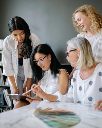 Vier Frauen arbeiten zusammen an einem Tisch, während sie auf ein Tablet schauen. Im Hintergrund sind Haftnotizen und Farbproben sichtbar.