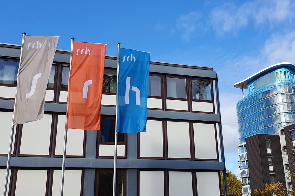 Fahnen in verschiedenen Farben mit dem Schriftzug "srh" vor einem modernen Gebäude. Im Hintergrund ist ein weiteres, gläsernes Gebäude sichtbar.