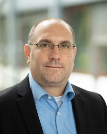 Ein Mann mit Brille trägt einen schwarzen Anzug und ein hellblaues Hemd. Er steht in einem modernen Innenraum mit unscharfem Hintergrund.