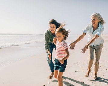 Drei Frauen, darunter ein Kind, laufen fröhlich am Strand entlang. Die Erwachsenen tragen lässige Kleidung, während das Kind ein gestreiftes T-Shirt und Shorts trägt. Die Wellen sind im Hintergrund sichtbar.